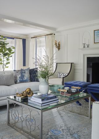 white living room with blue accents, glass coffee table, wainscoting, blue velvet stools, sofa, rug