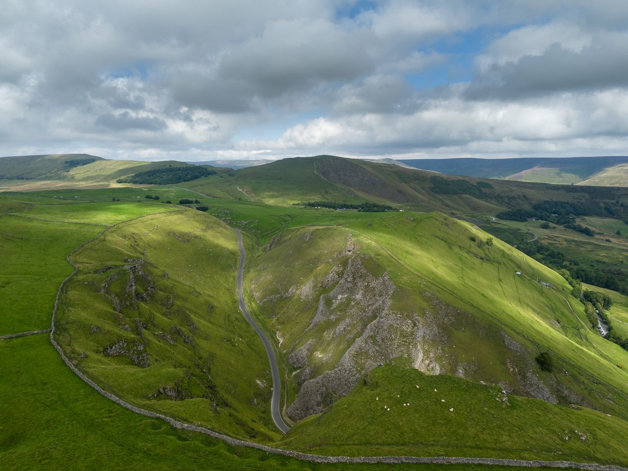 Image taken by DJI Air 3 (a camera drone). Here the focus is on a road winding through the green valleys. In the distance are rolling green hills dotted with dark green trees and a gray, cloudy sky.
