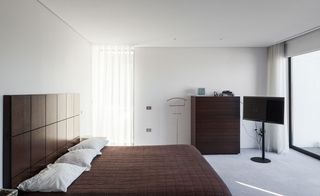 A bedroom with white walls, white ceiling and grey carpet. Brown chest of drawer positioned against the wall . A double bed with brown headboard, 3 white pillow cases and brown blanket