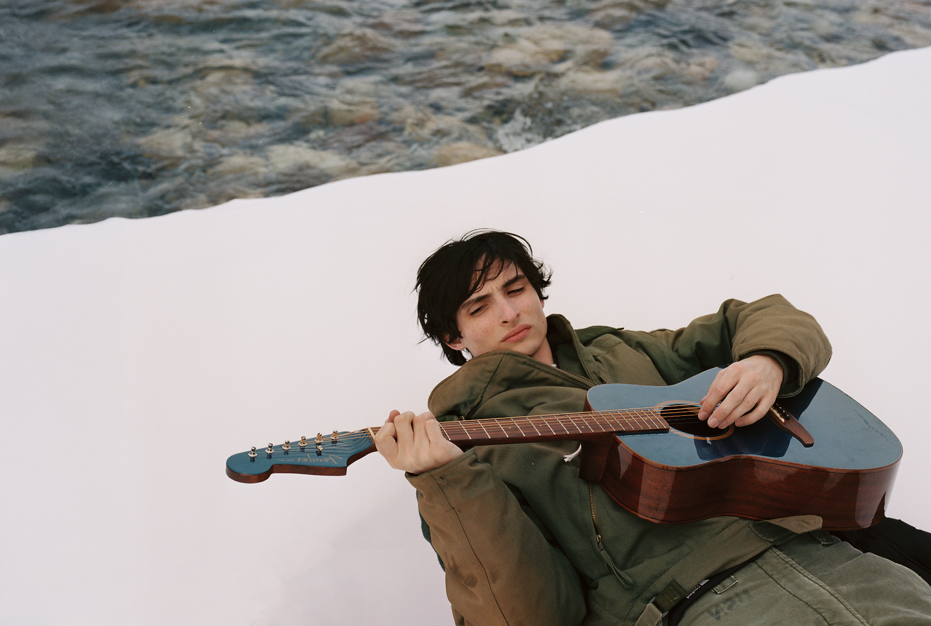 actor musician finn wolfhard lays in the show by a river holding an acoustic guitar in a press shot