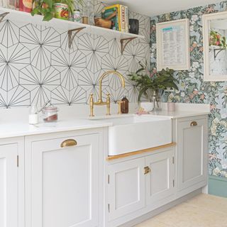 white kitchen with white units and worktop, patterned tile splash back, brass tap, open shelving, floral wallpaper and artwork, stone floor tiles