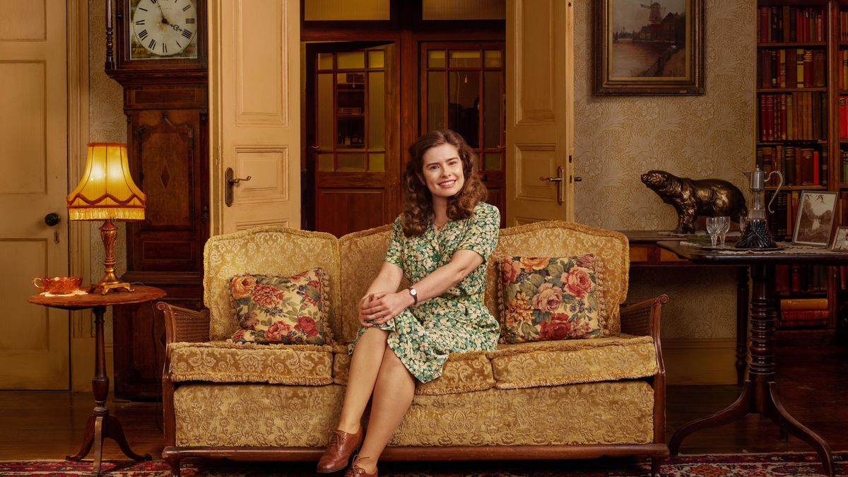 Rachel Shenton in a green floral dress as Helen sits on a sofa in Skeldale House in All Creatures Great and Small