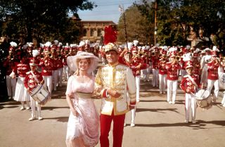 a marching band in red walks behind a band leader and a woman in a white dress in the movie adaptation of the music man