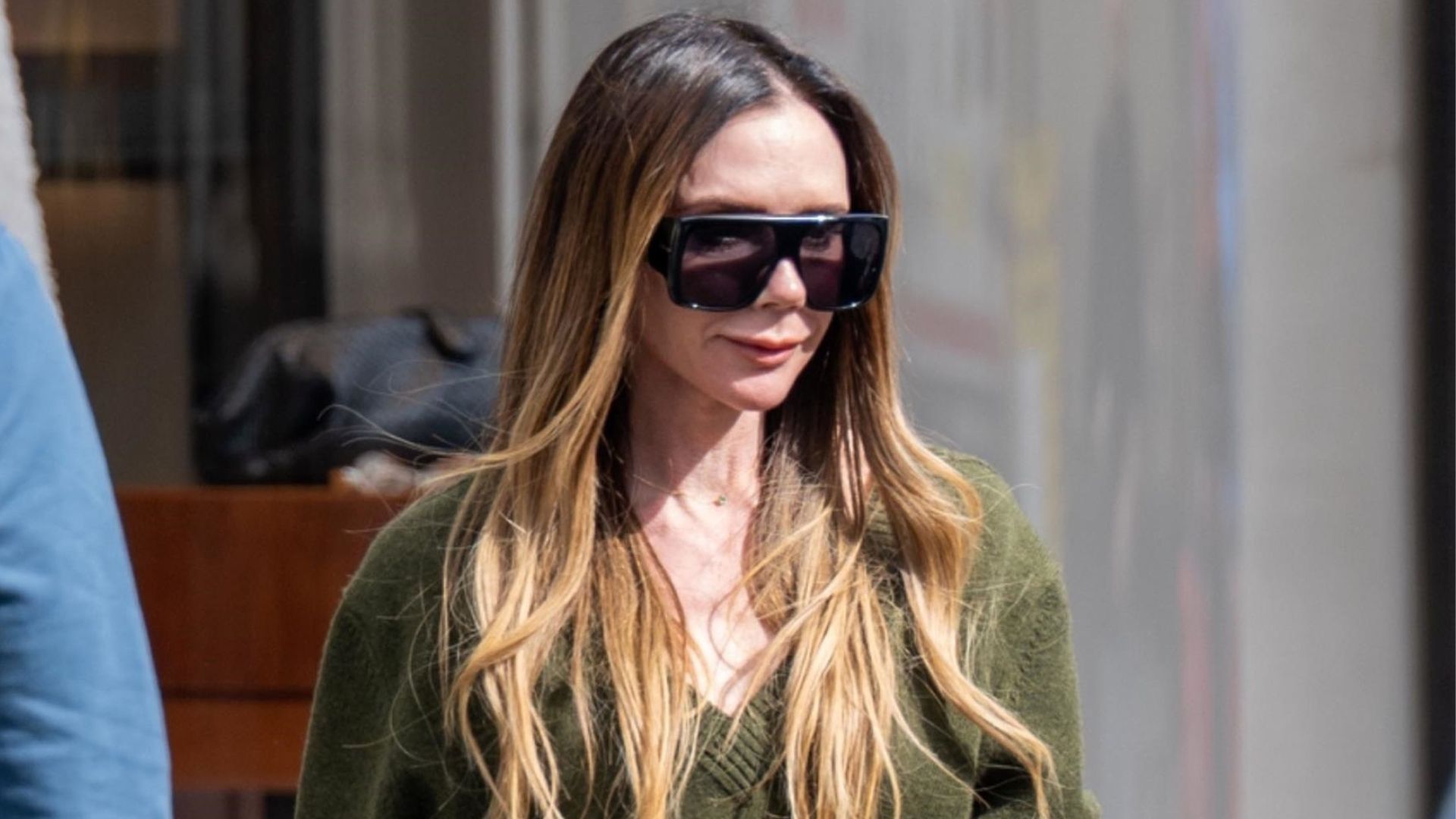 Victoria Beckham wears white jeans with an olive v-neck jumper and brown suede shoes.
