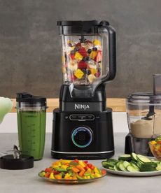 A Ninja blender filled with fruits, surrounded by cups of blended fruits and plates of cut vegetables, on a white counter against a gray background.