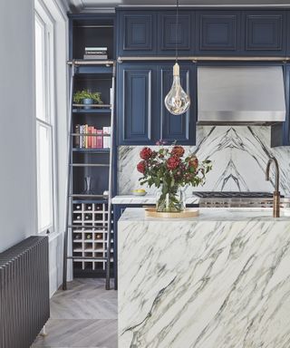 A kitchen island clad with white marble, with blue cabinetry on the rest of the kitchen, wine rack and ladder to top shelves