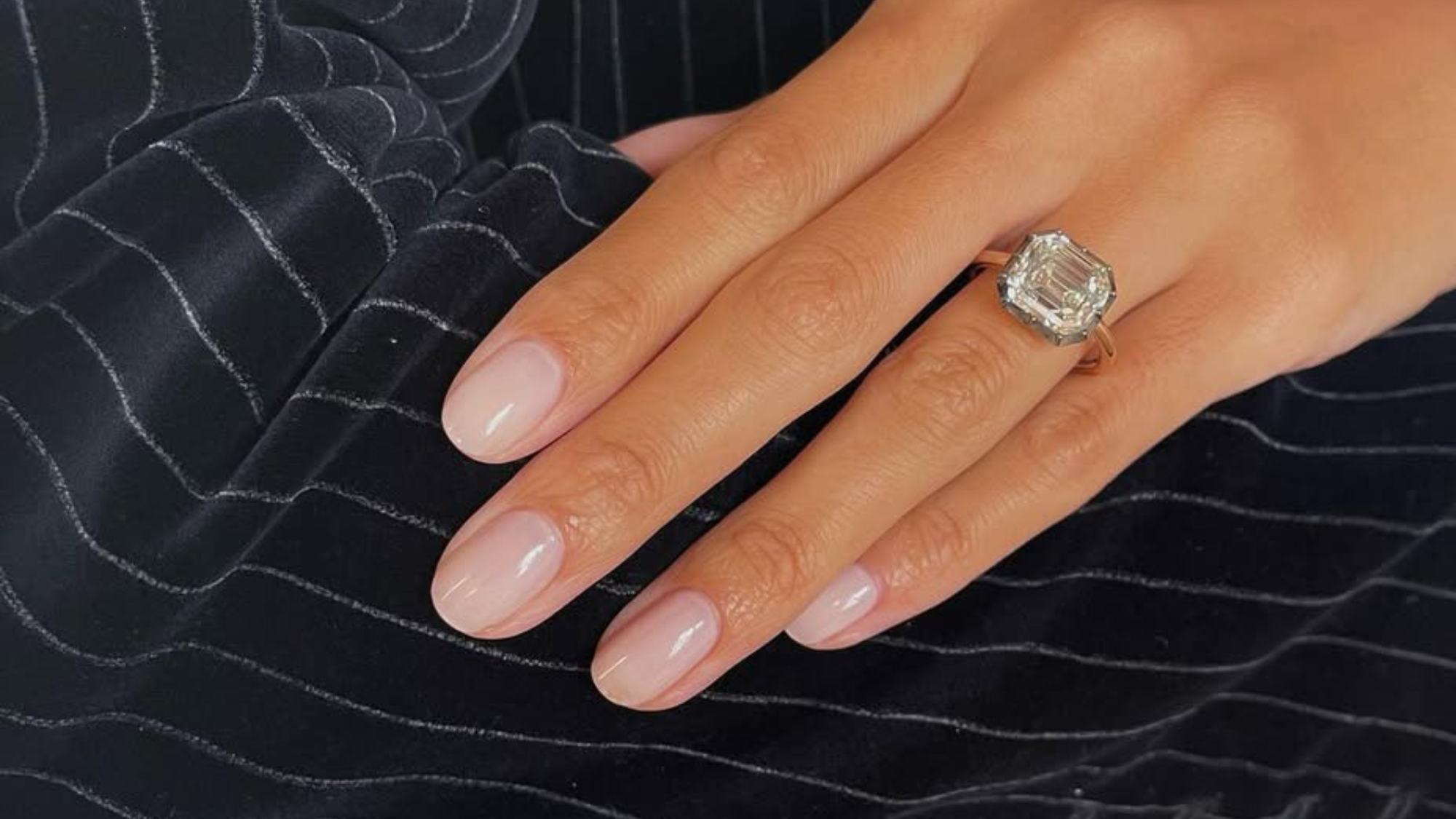 Graceful hand with a barely there, pinky nude manicure, radiant cut diamond ring, resting on an arm in a pinstripe jacket