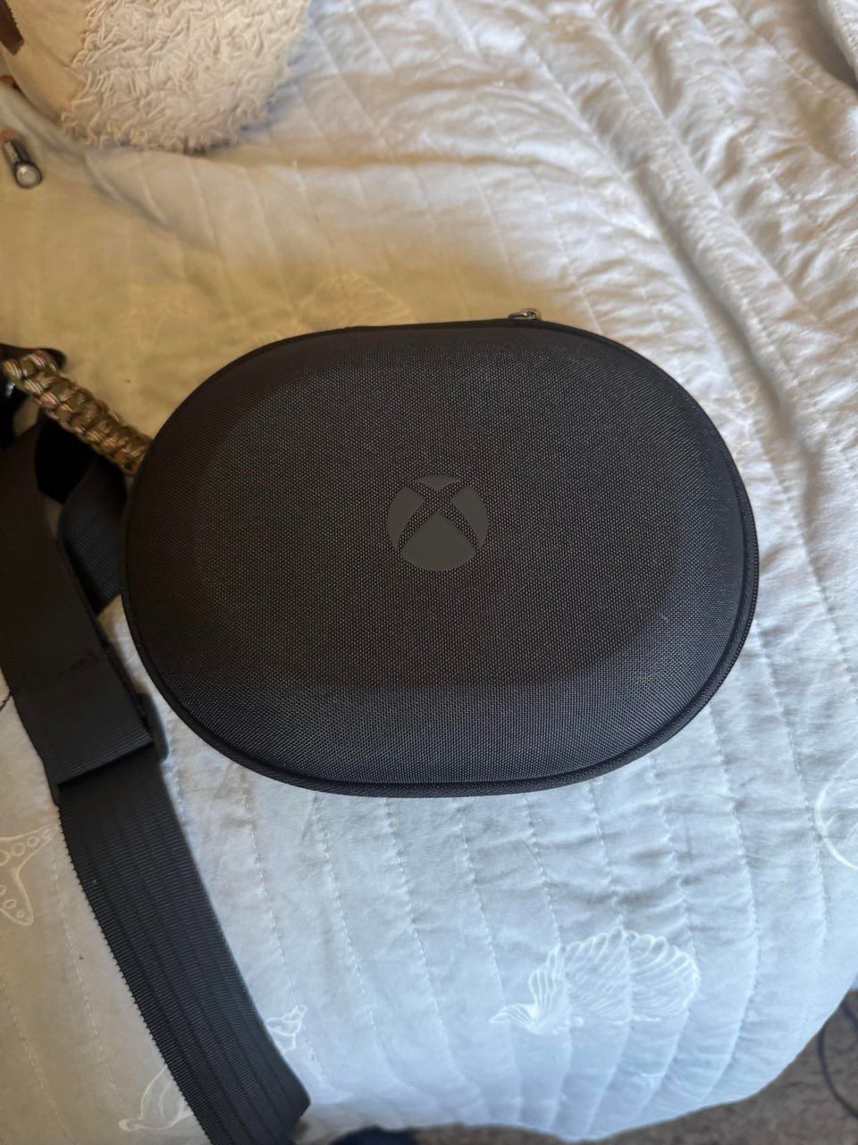 A black, oval-shaped, hardshell carrying case for an Xbox controller with the Xbox logo centered on the lid, resting on a white quilted blanket.