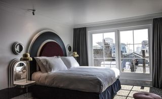 A room in the Henrietta Hotel. A large bed with white linen and a rounded headboard in deep red and black is to the left, next to which is a night table with a golden lamp. To the far wall, there is a double french style window, overlooking the roofs of other buildings.