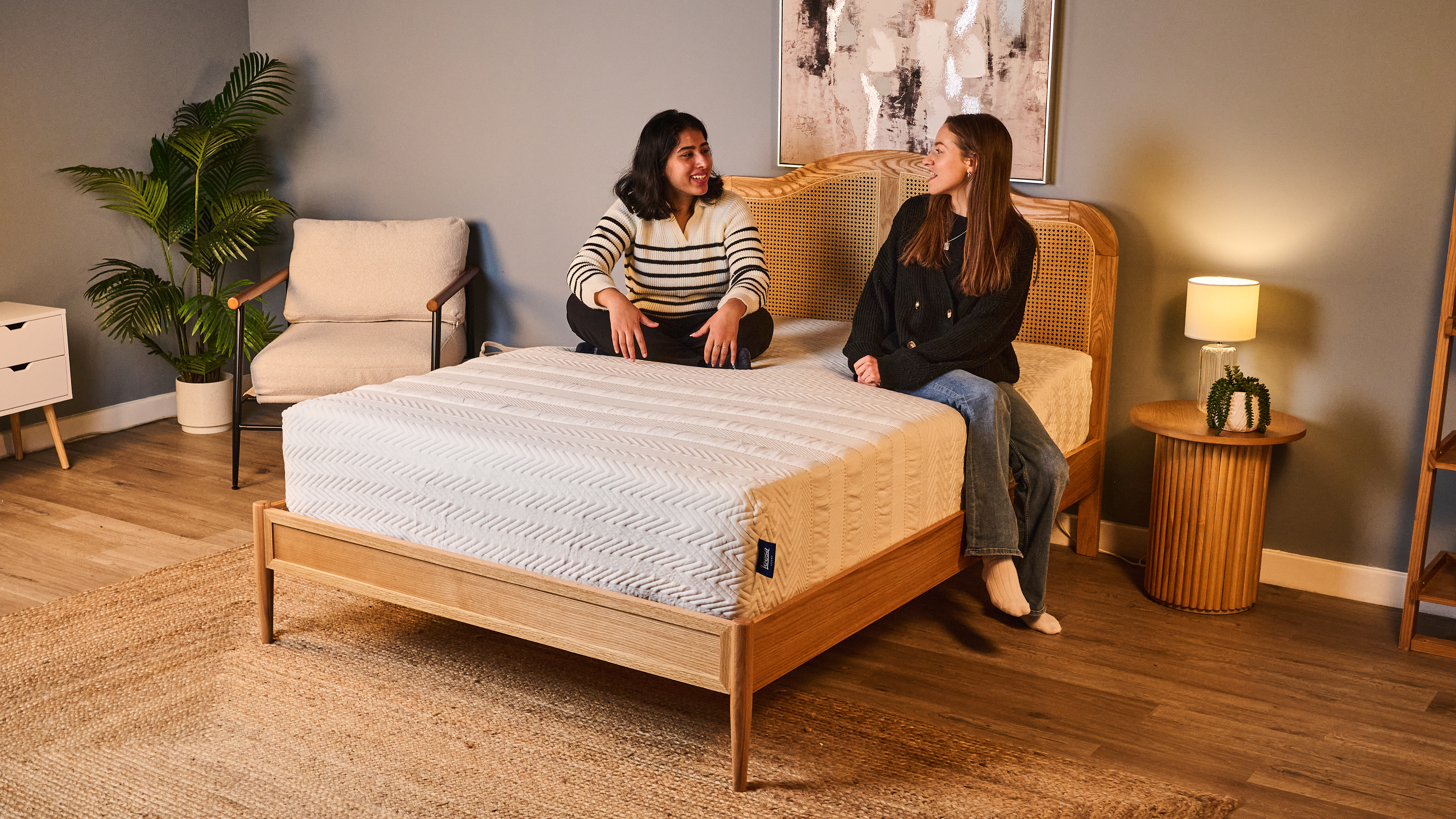 Two women sit on the Leesa Legend Hybrid Mattress in a light blue warmly-lit bedroom.