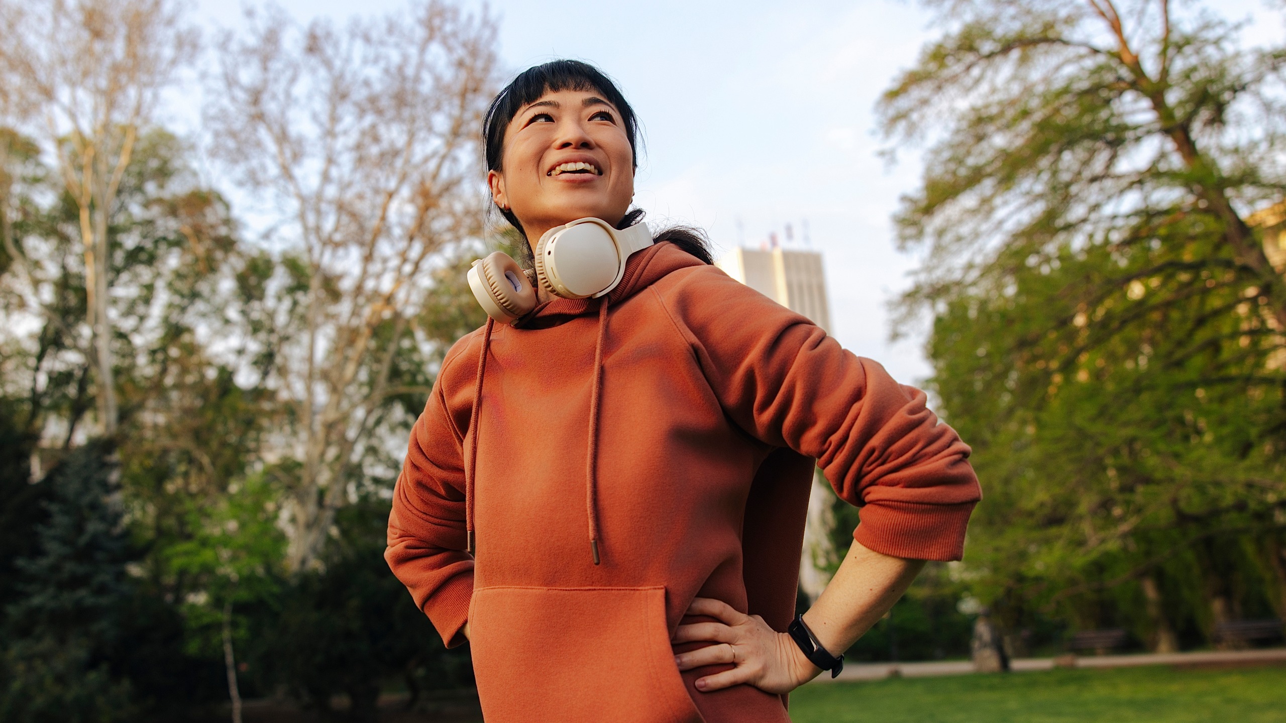 A woman in a park wearing headphones stands looking at the sky with her hands on her hips.