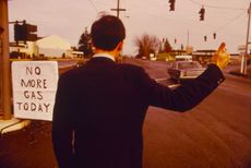 A man in a suit stands at a gas station, trying to hitchhike in the 1970s.