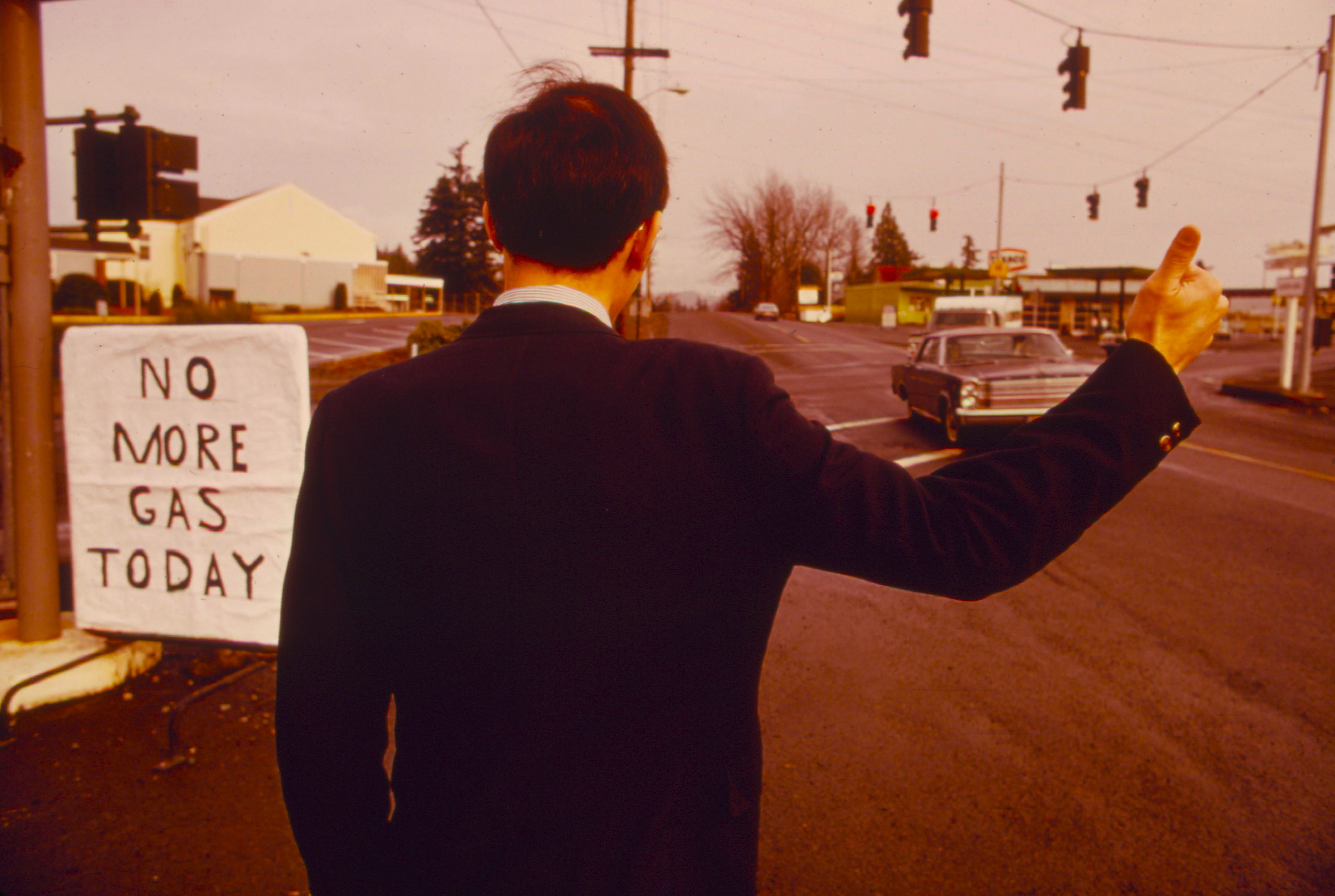 A man in a suit stands at a gas station, trying to hitchhike in the 1970s.