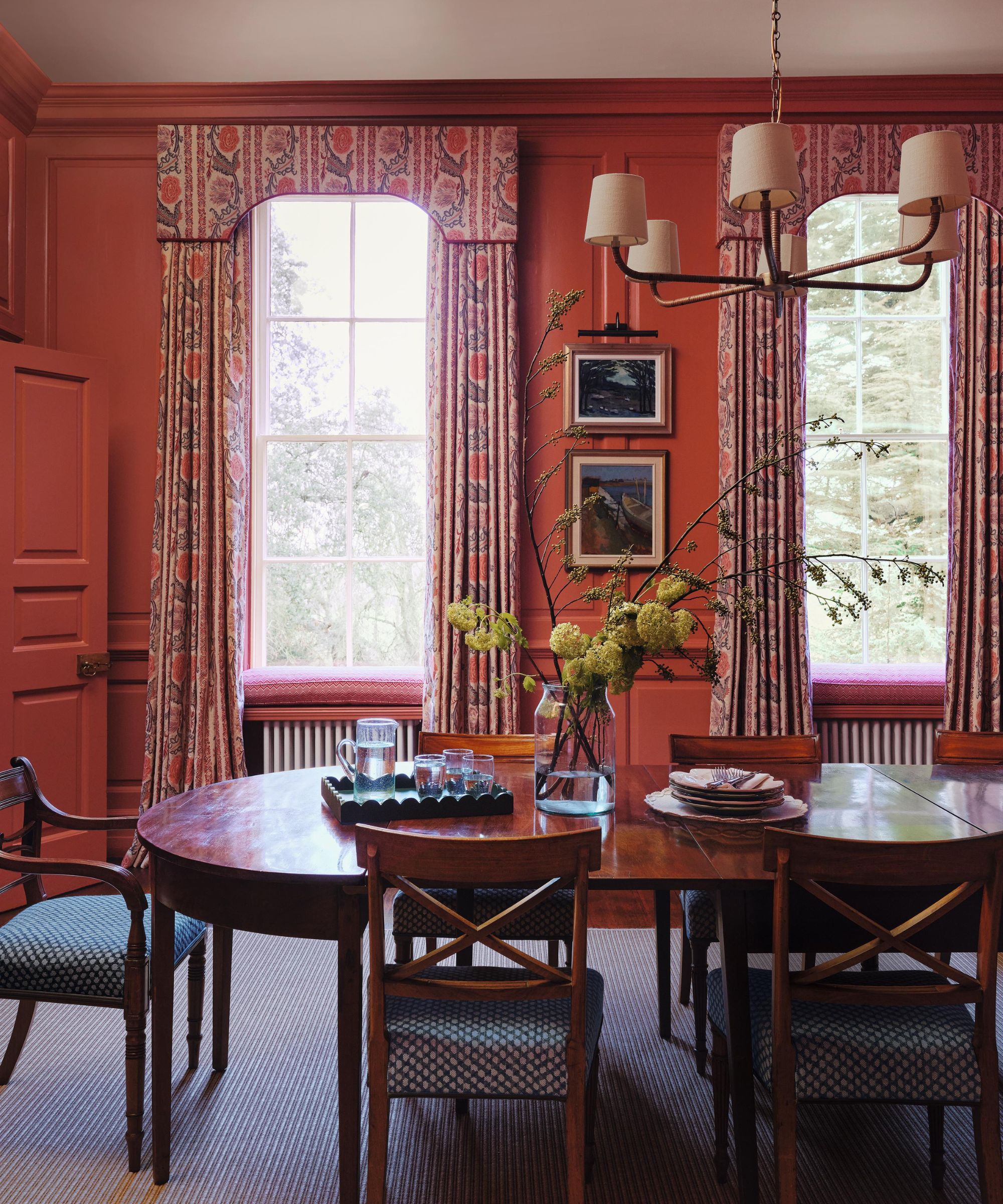 a traditional English dining room with paneled walls painted red with floral curtains with pelmets and an antique wood dining table and chairs