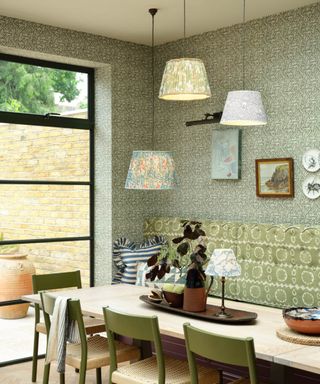 series of 3 fabric pleated patterned light shades hung at different heights over wooden dining table, green and wicker dining chairs, green and white patterned wallpaper on walls and large crittall style floor to ceiling windows to side