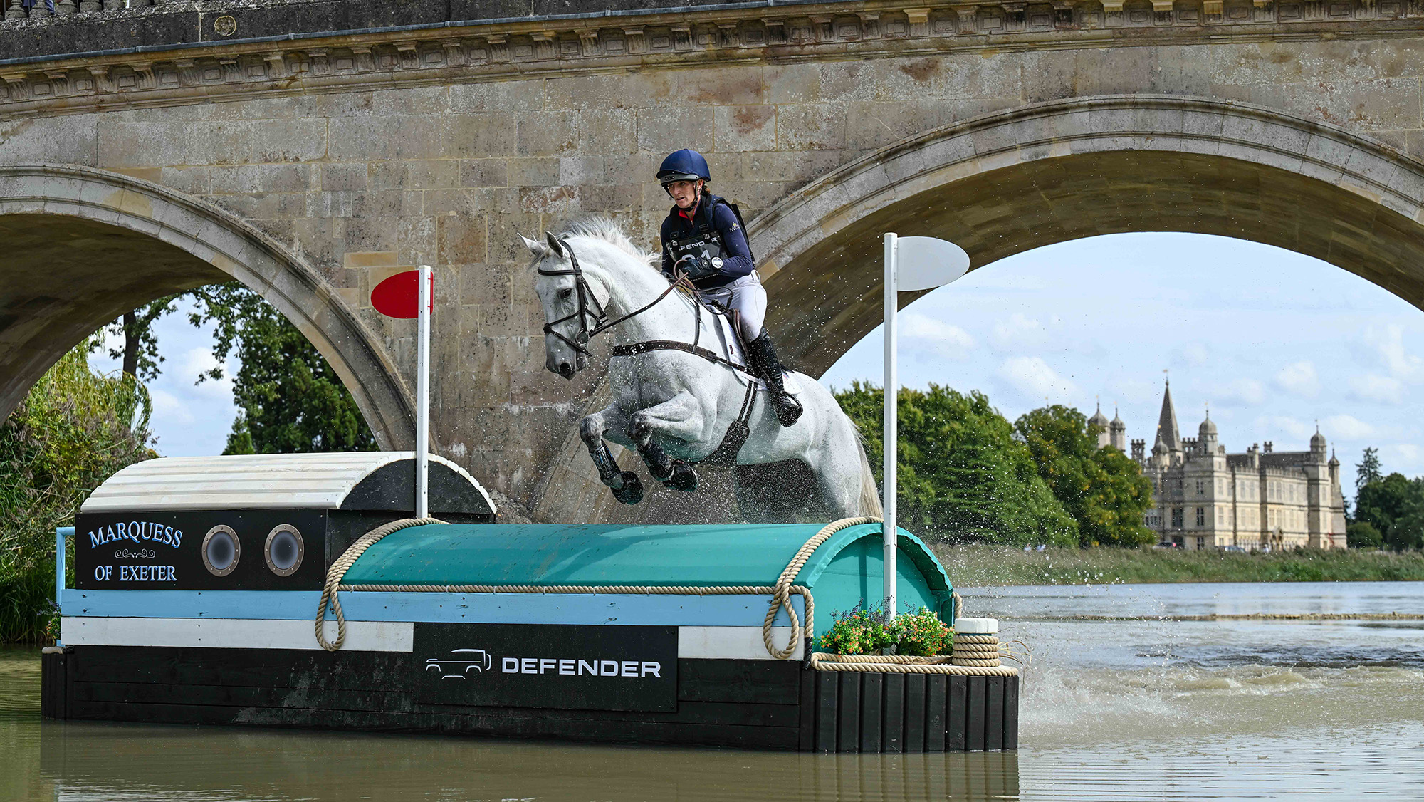 Holly Richardson, a rider in a blue top and hat cover, and Ballyneety Silver Service, a grey horse, jump a boat fence in water at Burghley 2025