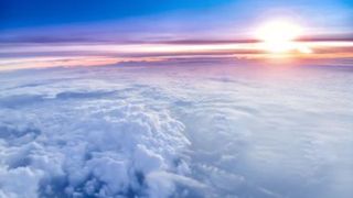 an aerial view of the sun setting above a large bank of clouds. layers of color in the sky show a fading from bright orange above the cloud bank to dark blue at the top of the picture