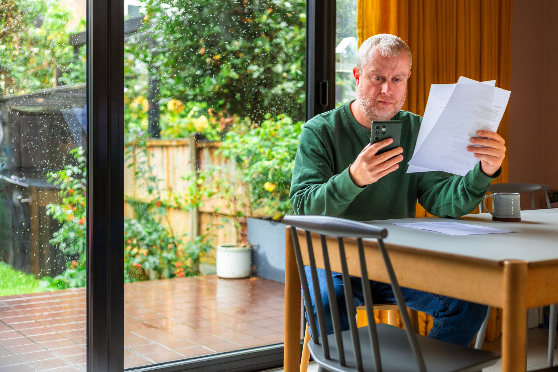 Portrait of a mid adult man checking his bills at home, using his smart phone. He is dressed casually in a green crewneck sweater and jeans while in his modern kitchen.