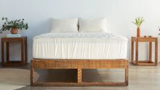 White Avocado mattress protector on a wooden bedframe in a white minimalist bedroom, with wooden bedside tables to either side with houseplants, wooden flooring, white walls, and a circular cream rug.