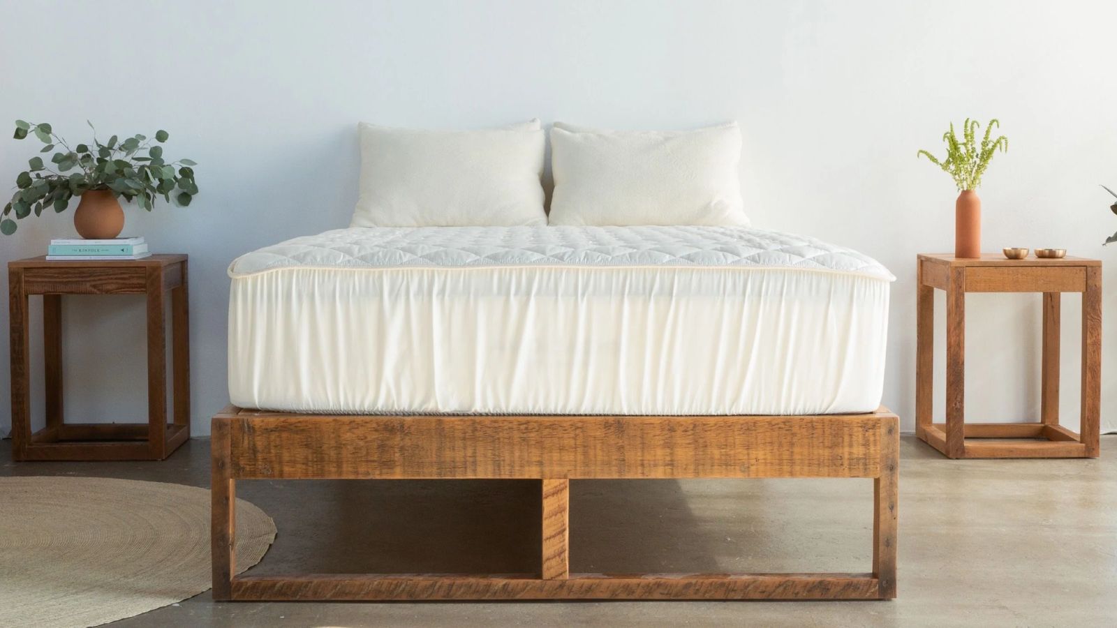 White Avocado mattress protector on a wooden bedframe in a white minimalist bedroom, with wooden bedside tables to either side with houseplants, wooden flooring, white walls, and a circular cream rug.