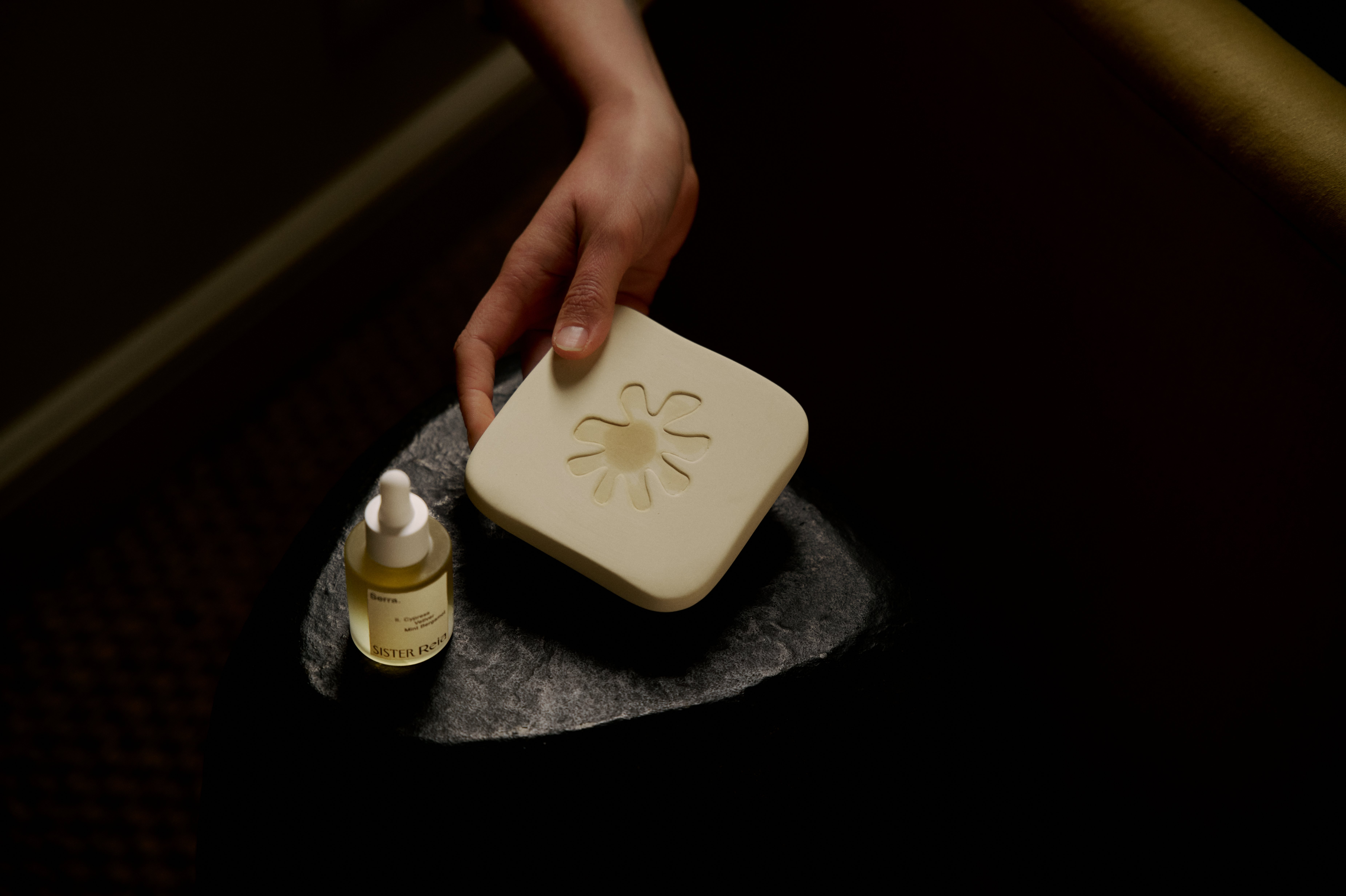 A room lit by a sunset boasts a home fragrance diffuser in the form of a pale yellow box, with a bottle of perfume, a vase of flowers, books, and other collectibles like candles and home accessories.