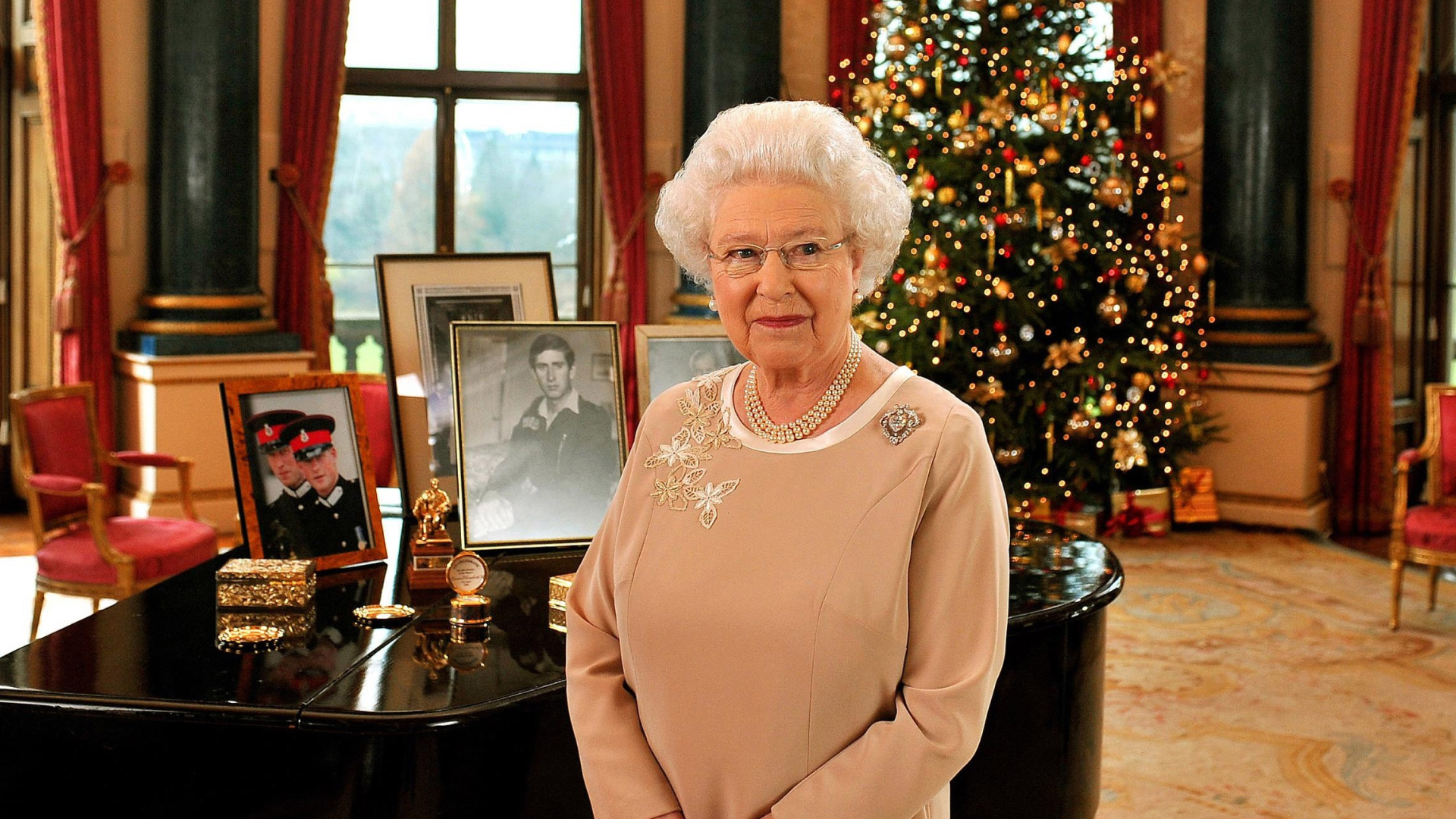 Queen Elizabeth in a tan dress standing in front of a piano and christmas tree