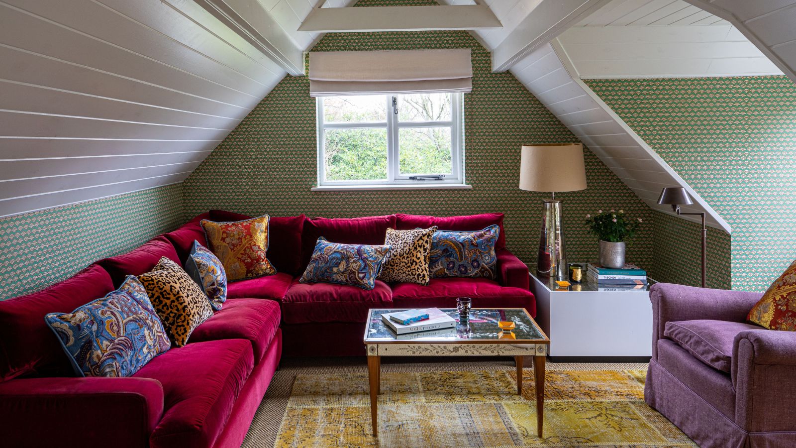 wallpapered snug room set in a loft or attic space with a pitched panelled ceiling and green wallpaper with a large pink velvet couch 