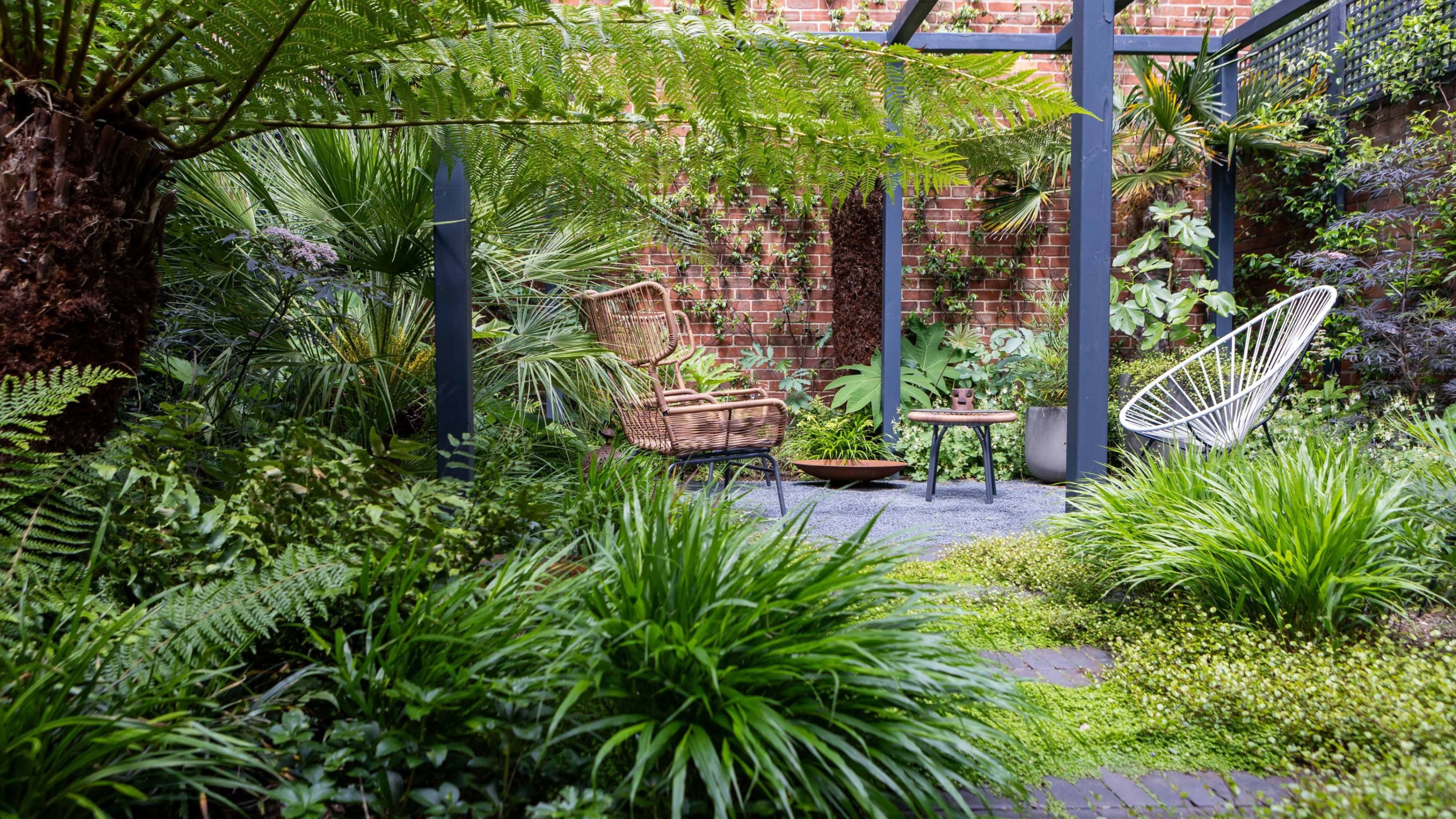 A calming brick-walled urban garden heavily covered in green foliage with repeat planting and a pergola by GRDN