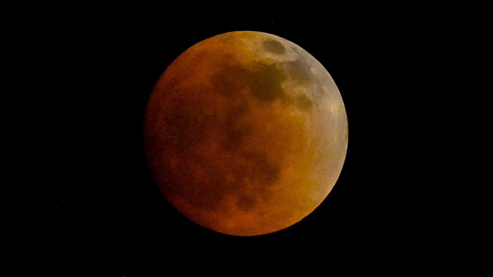 A red-orange full moon is shown hanging against a black sky, with dark basaltic plains called lunar maria marking its surface. The left side of the moon is slightly darker than the right.
