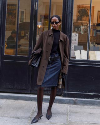 French influencer wearing black turleneck sweater, denim midi skirt, brown coats, tights, and black heeled mules.