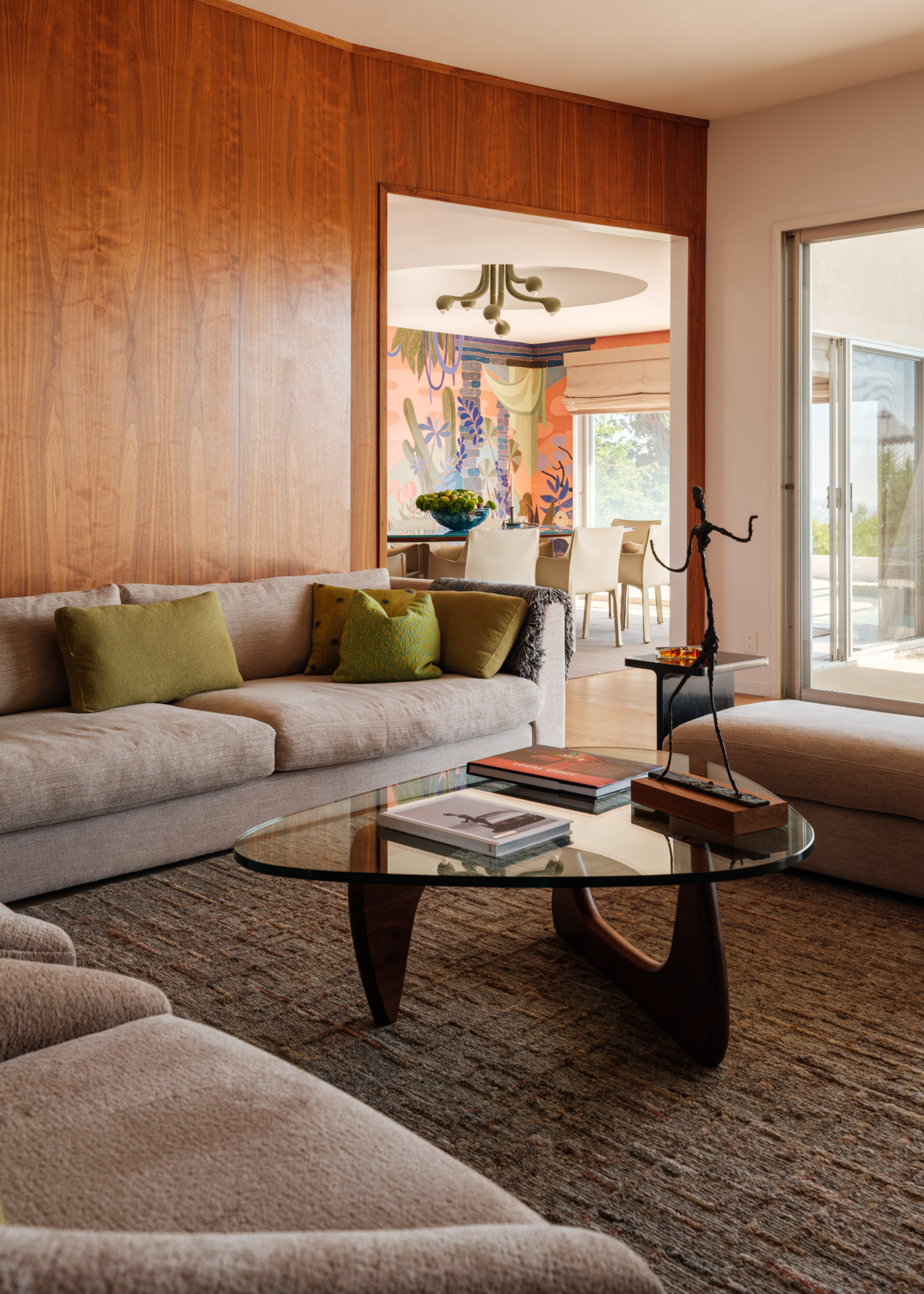 A wood living room with a gray couch with green cushions, a glass coffee table with books and a metal statue, and a textured area rug
