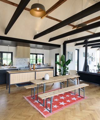 large kitchen diner with vaulted ceiling, pale wooden cabinets and bare wood dining table with metal legs and matching benches with red rug with white stars underneath