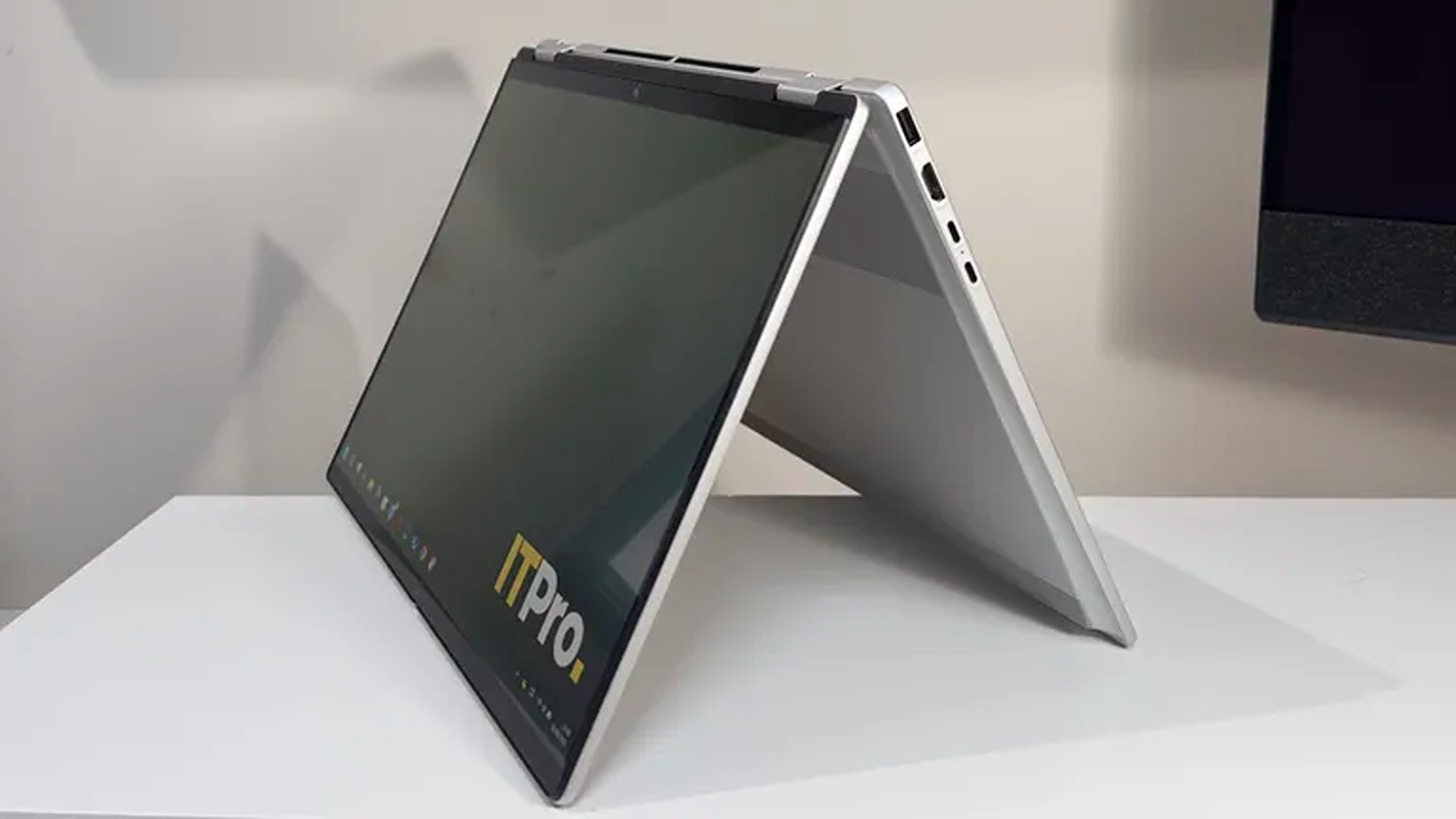 A silver 2-in-1 laptop folds into a tent-like stand on a white table, its dark touchscreen showing an ITPro logo; clean, modern product display.