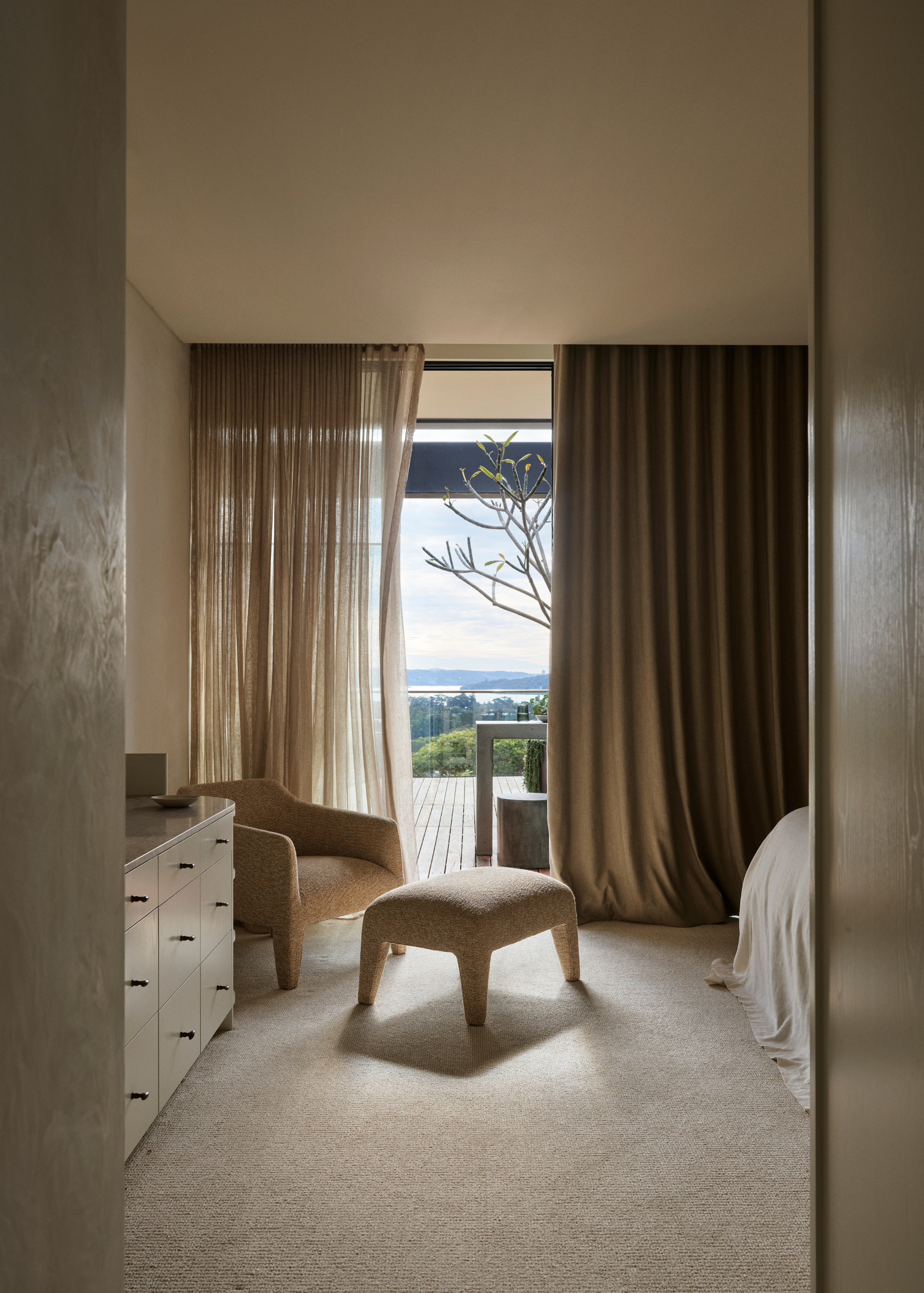 A bedroom with an attached balcony featuring layered curtains, a soft sofa chair and pouf, and a chest of drawers