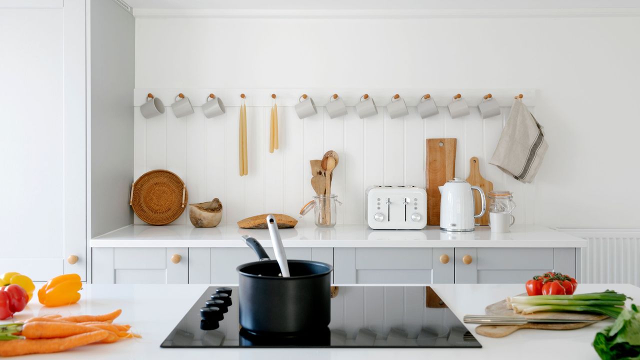 White kitchen with white worktops, including a black electric hob with a saucepan on it surrounded by vegetables