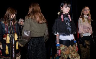 Four female models wearing looks from the Dsquared2 collection. One model is wearing a black coat with multicoloured detail and red and yellow tassels. Next to her is a model wearing a army green coloured ribbed piece with brown leather elbow patches and black bottoms. The third model is wearing a white top with a black lace cape style overlay, a black buttoned belt, brown and green camouflage style trousers and various accessories in different colours. And the fourth model is wearing a light coloured top with a cream lace overlay, army green trousers, pink tassel earrings and black gloves. There is a fifth model in the background with short blonde hair