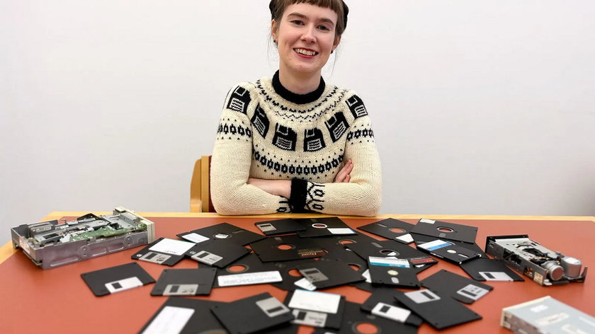 A smiling woman with her arms folded sits at a table scattered with floppy disks. 