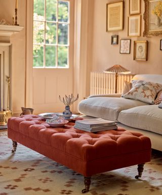 A cream-painted living, naturally lit, with a large white soft sofa and a large red padded ottoman with books piled on top