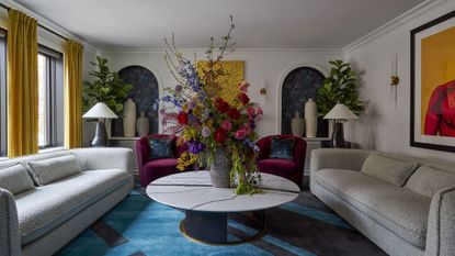 A white living room with yellow curtains, potted fiddle leaf figs, a dramatic floral arrangement on a stone coffee table, a blue patterned area rug, purple sofa chairs, gray sofas and ceramic vases