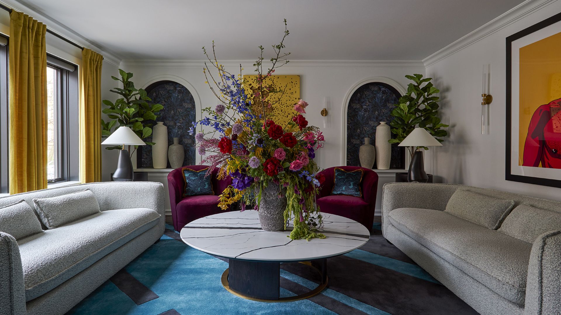 A white living room with yellow curtains, potted fiddle leaf figs, a dramatic floral arrangement on a stone coffee table, a blue patterned area rug, purple sofa chairs, gray sofas and ceramic vases