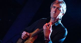 Allan Holdsworth frets a chord on his headless electric guitar, onstage in Spain, 2006. A white stage light illuminates the right-hand side of his face.