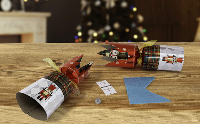 An open Christmas cracker shows a single dice and a paper crown on a wooden table. A Christmas tree is lit up in the distance. 