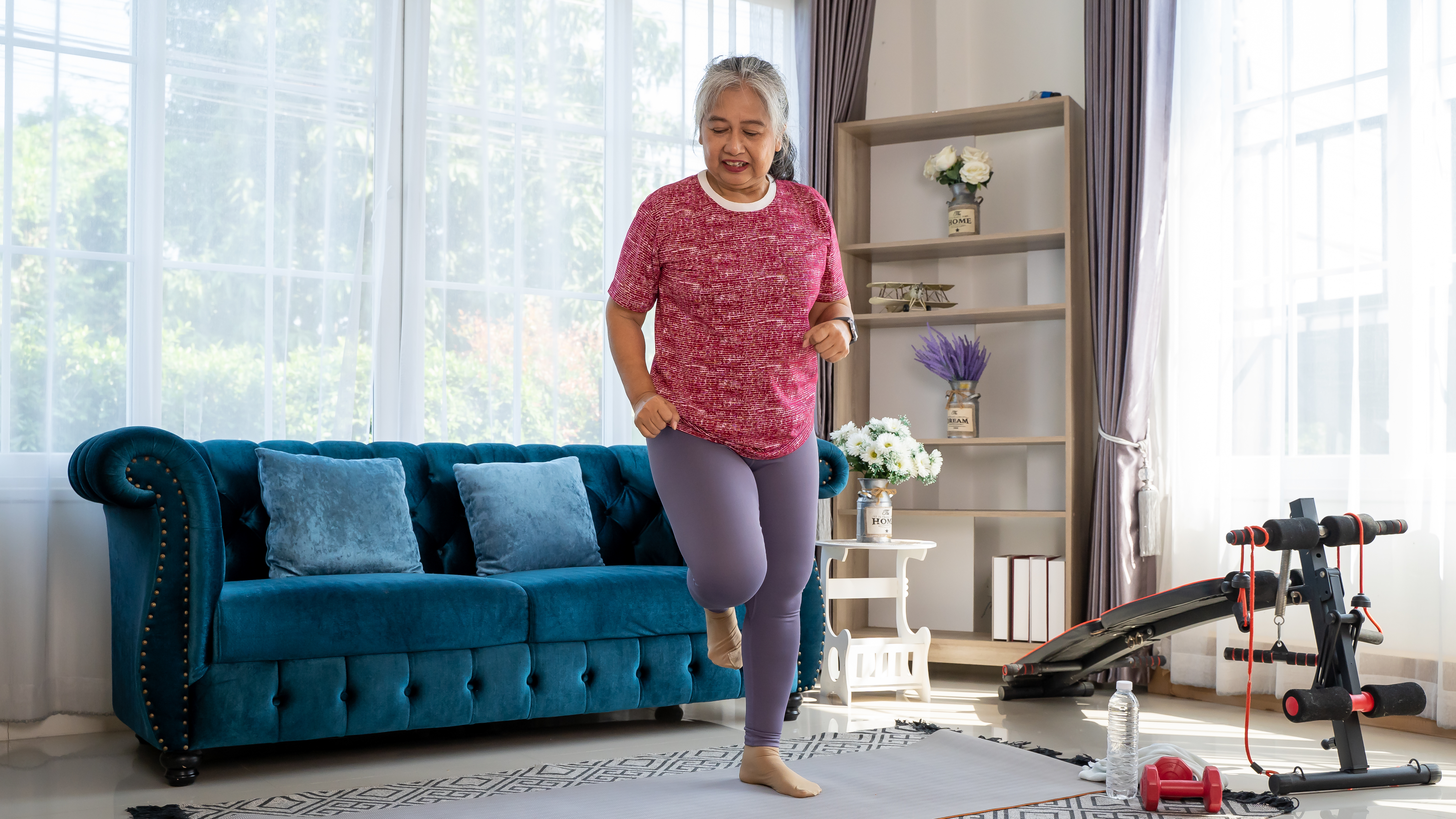 A woman exercises at home, jumping onto one leg. She's in her living room in front of a couch, with exercise equipment next to her.