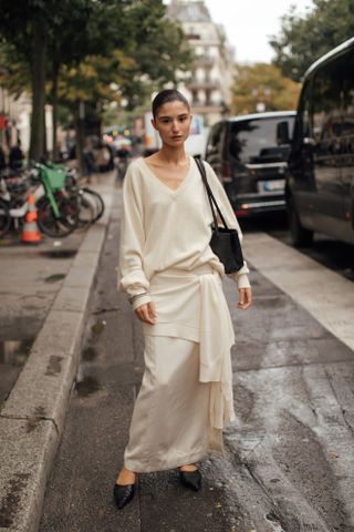 A woman standing in the street at Milan Fashion Week in a white slip skirt, white sweater, black shoes, and with a white sweater tied around her waist.