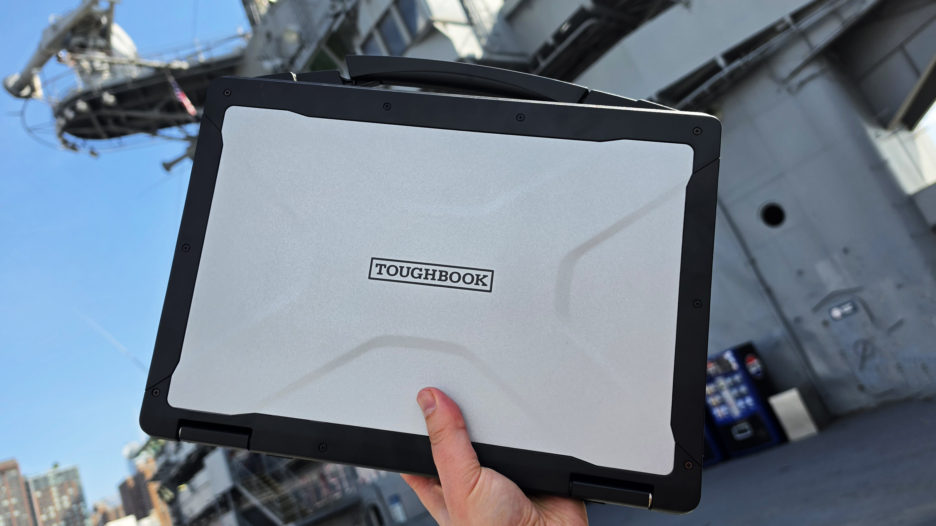 A hand holds a rugged Toughbook laptop outdoors against a backdrop of an industrial or naval setting. The bright blue sky adds contrast, emphasizing durability.