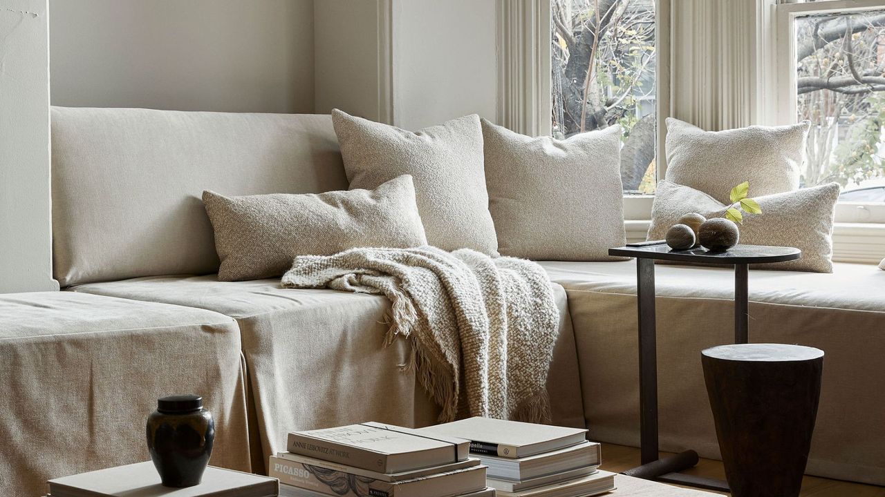 A cozy white living room with layered throws, pillows, and stacks of books with vases