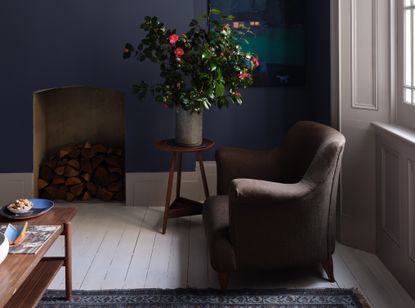 A living room painted dark blue with an armchair by the window and logs stacked in the fireplace