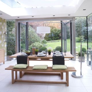 A modern glass garden conservatory with wooden dining table, wooden benches, green cushions and folding doors