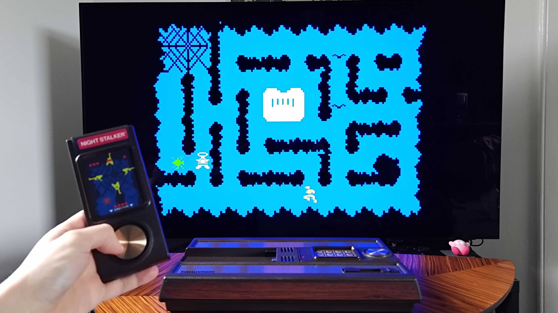 Hand holding Intellivision Sprint controller with Night Stalker gameplay on TV in backdrop.