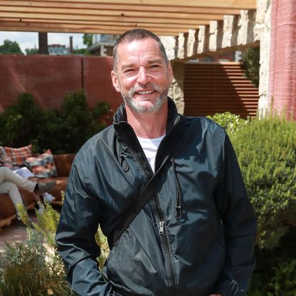 Fred Sirieix in a black zip top in front of a green hedge and wooden pergola